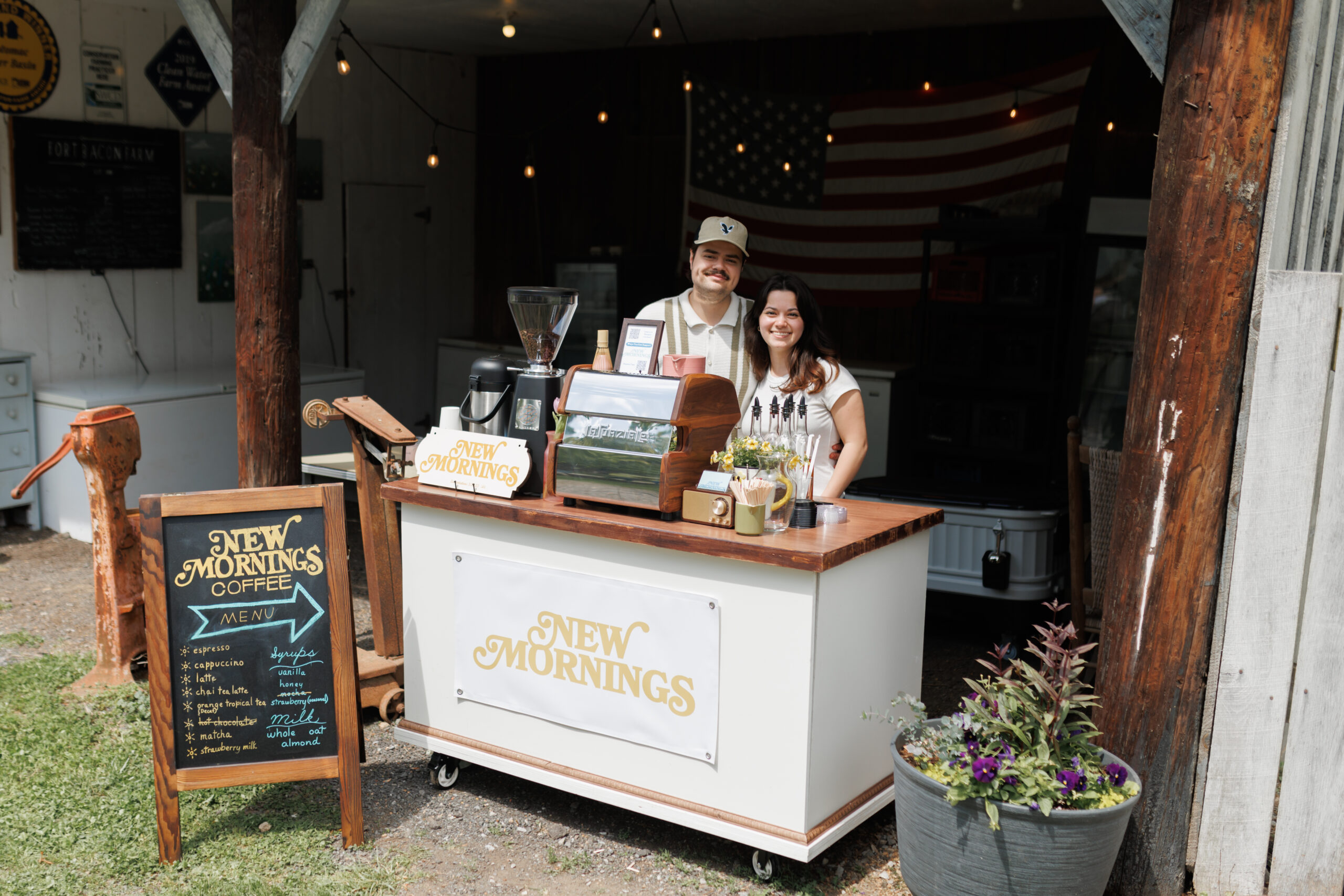 Owners of New Mornings Coffee standing behind their mobile espresso bar with a handwritten menu outside a rustic barn venue