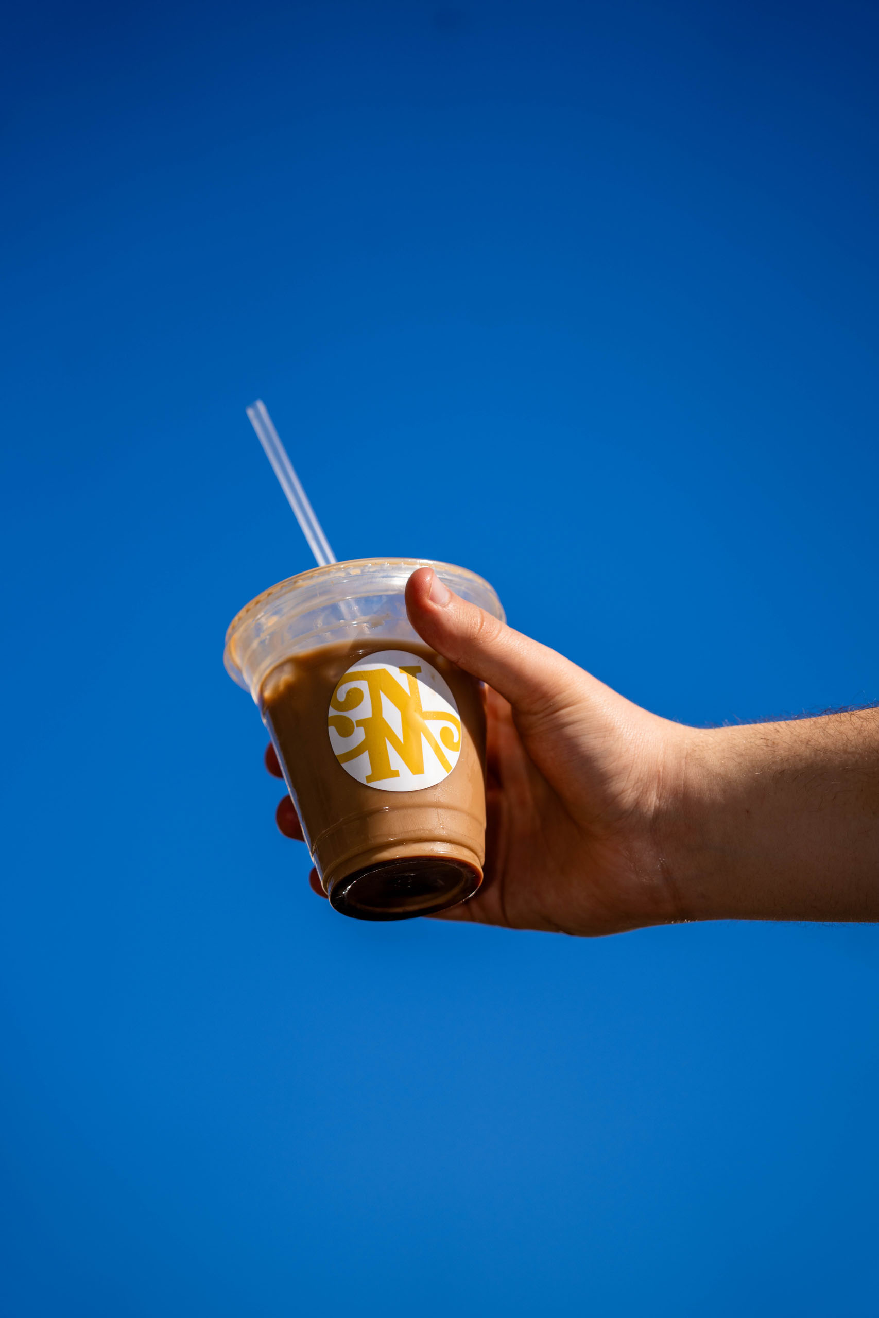 A hand holding a New Mornings Coffee iced latte against a clear blue sky, featuring the brand's signature gold monogram logo
